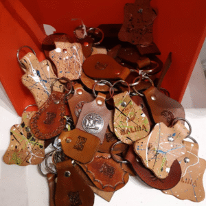 A collection of handmade leather keychains in various shapes, colors, and designs piled inside a red container. Some keychains feature embossed initials, others are decorated with paint splatters, engraved patterns, or metal emblems.