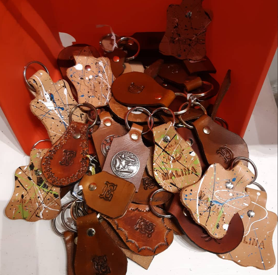A collection of handmade leather keychains in various shapes, colors, and designs piled inside a red container. Some keychains feature embossed initials, others are decorated with paint splatters, engraved patterns, or metal emblems.